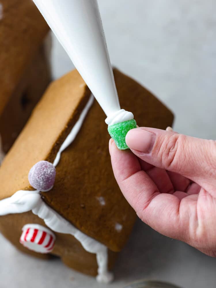 Nothing says holiday season like a Gingerbread house sitting on the counter! This recipe includes the recipe for gingerbread, a template to print out the ginger bread house pieces, and how to make the royal icing. This is the perfect recipe for the whole family to enjoy! A hand piping royal icing onto the bottom of gumdrops with a gingerbread house in the background.