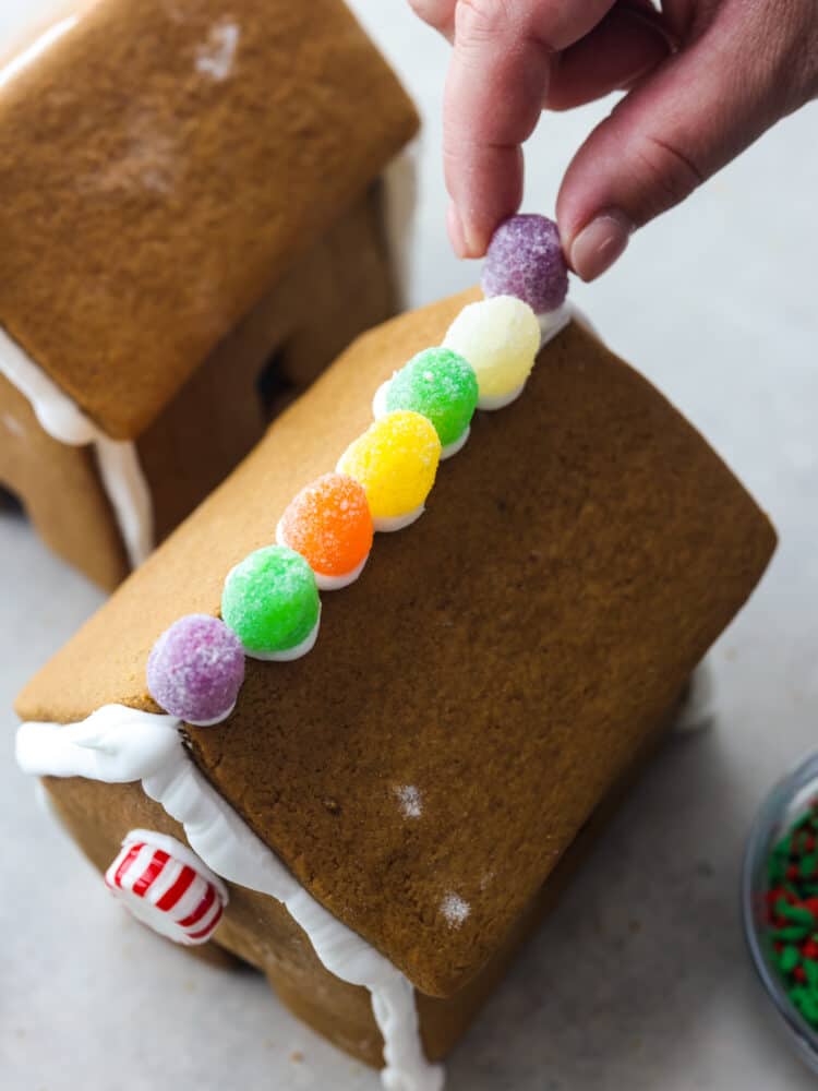 Nothing says holiday season like a Gingerbread house sitting on the counter! This recipe includes the recipe for gingerbread, a template to print out the ginger bread house pieces, and how to make the royal icing. This is the perfect recipe for the whole family to enjoy! A hand placing the gum drops on top of the roof of a gingerbread house.