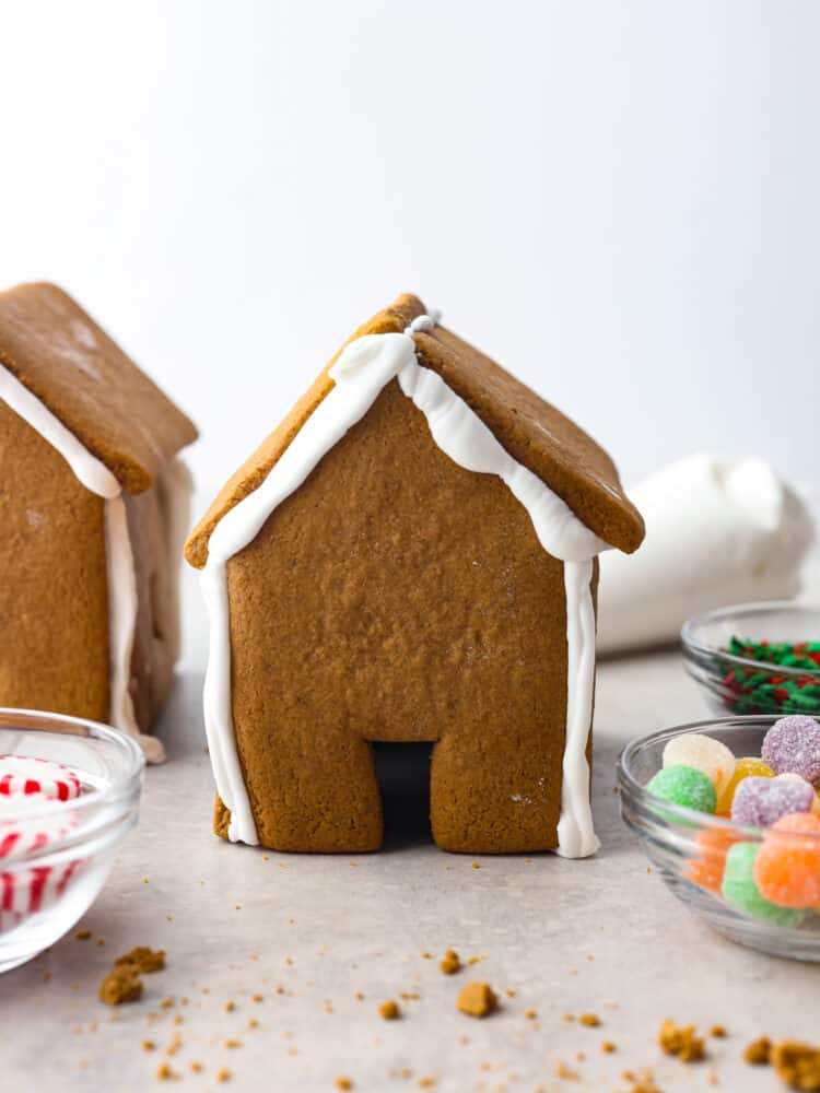 Nothing says holiday season like a Gingerbread house sitting on the counter! This recipe includes the recipe for gingerbread, a template to print out the ginger bread house pieces, and how to make the royal icing. This is the perfect recipe for the whole family to enjoy! Gingerbread house with bowls of candy around it and another house in the background.