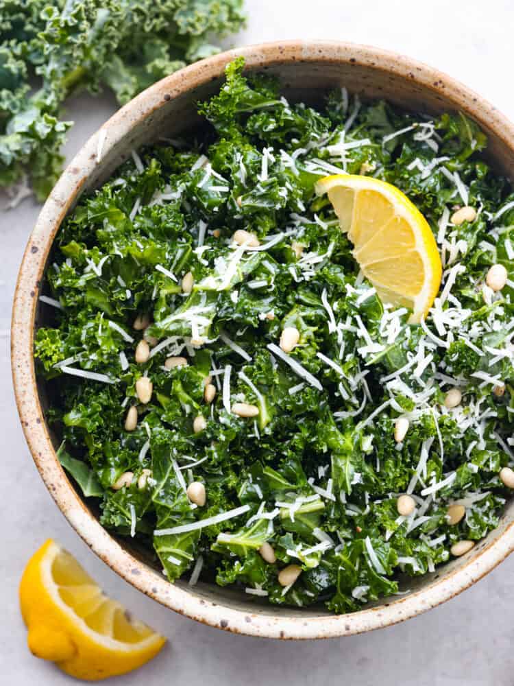 If youโre looking for a tasty way to incorporate more leafy greens into your diet, this is it! This kale salad is tossed with parmesan, lemon, and pine nuts for a bright and fresh flavor combination you won't be able to resist! Top-down view of kale salad in a brown serving bowl, garnished with a lemon.