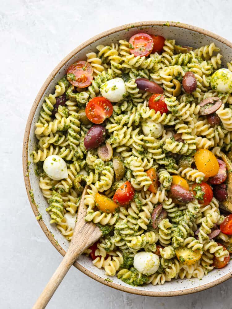 This pesto pasta salad is loaded up with fresh mozzarella, and juicy tomatoes, coated with an easy-to-make homemade pesto sauce made with fresh basil and garlic. It's so fresh and delicious! The top view of a bowl with pesto pasta salade and a wooden spoon in it.