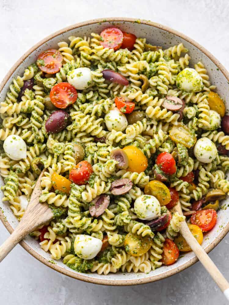 This pesto pasta salad is loaded up with fresh mozzarella, and juicy tomatoes, coated with an easy-to-make homemade pesto sauce made with fresh basil and garlic. It's so fresh and delicious! The top view of a bowl of pasta salad with 2 wooden spoons.
