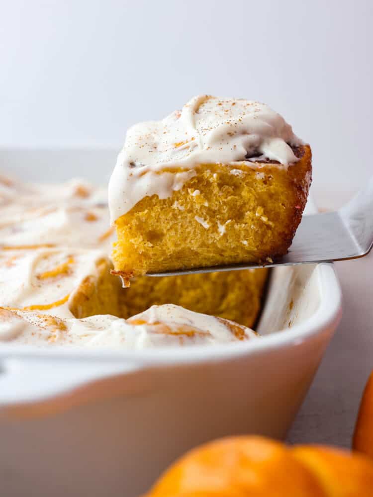 Pumpkin cinnamon rolls are filled with a delicious brown sugar filling and smothered in homemade cream cheese frosting. Soft, fluffy and so perfect for fall! A cinnamon roll being taken out of a white pan with a metal spatula.