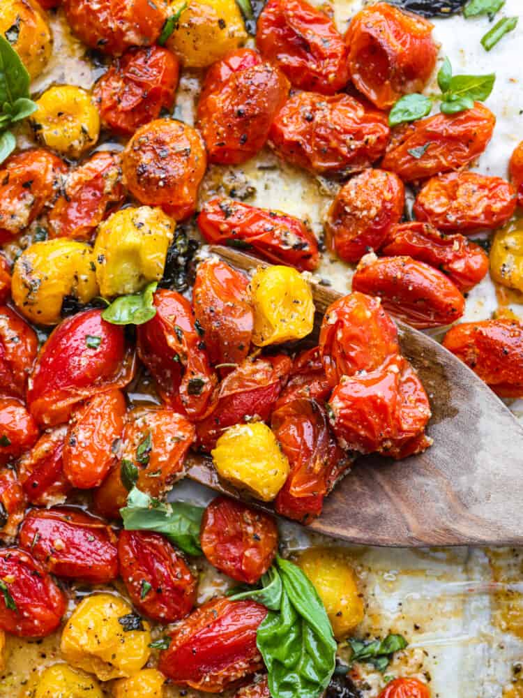 These roasted cherry tomatoes are seasoned with garlic and basil and literally bursting to perfection. It's a simple and delicious side dish that you will want to pair with every meal! A close up of roasted cherry tomatoes being scooped up with a wooden spoon.