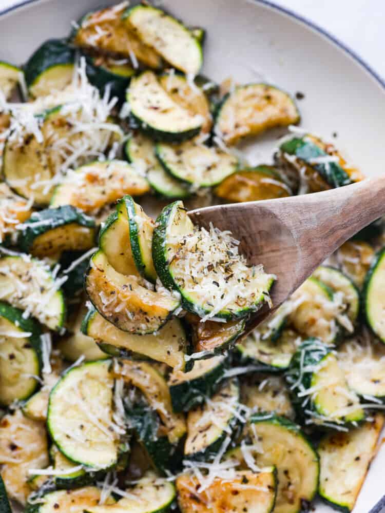 Sautéed zucchini is a fresh and simple side dish bursting with cheesy Italian flavors. Garden zucchini is sautéed in olive oil and fresh garlic, perfectly seasoned with Italian herbs, and then topped with parmesan cheese. A close up photo of a wood serving spoon lifting the sautéed zucchini from the skillet.