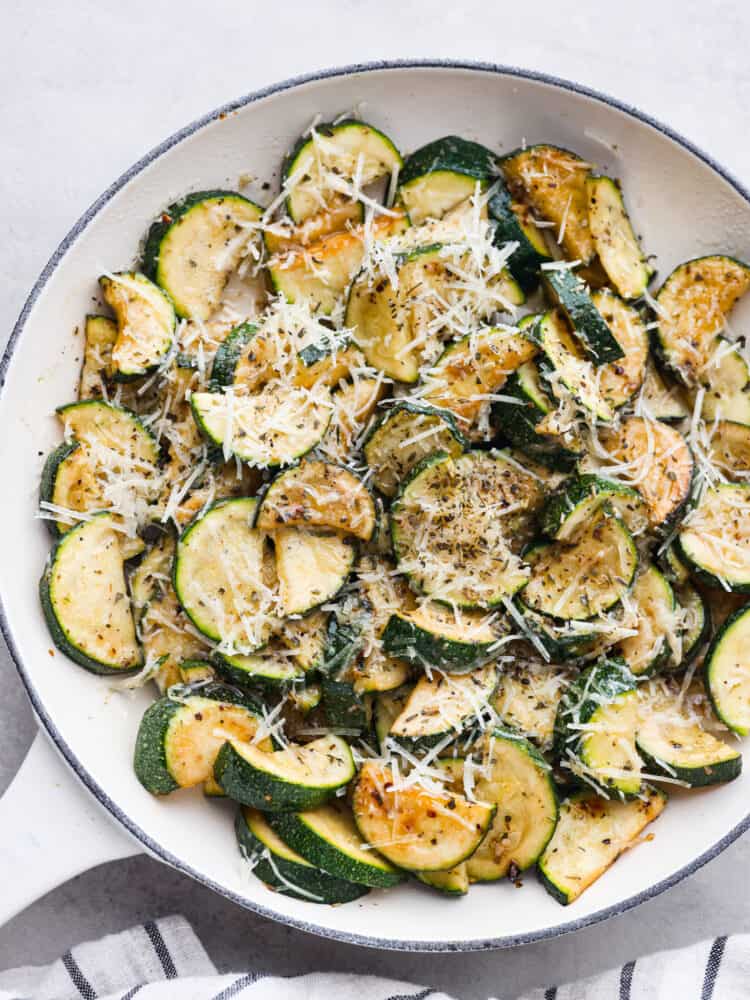 Sautéed zucchini is a fresh and simple side dish bursting with cheesy Italian flavors. Garden zucchini is sautéed in olive oil and fresh garlic, perfectly seasoned with Italian herbs, and then topped with parmesan cheese. Top view of sautéed zucchini and melted parmesan cheese on top. The sautéed zucchini is beautifully displayed in a large white skillet with a striped towel next to it.