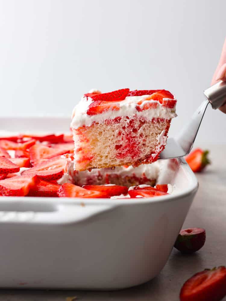 Incredibly light and tasty, this strawberry poke cake will be the star of any gathering! It's airy, soft, and brings that fresh strawberry taste into every single bite! The side view of a slice of strawberry poke cake being taken out of the pan with a silver cake server.