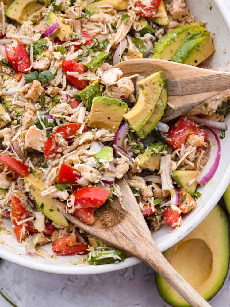This fresh and flavorful avocado tuna salad is the perfect side dish for summer! The combo of bright veggies, savory tuna, and creamy avocado is one you don't want to miss! A bowl of tuna salad being mixed with two wooden spoons.