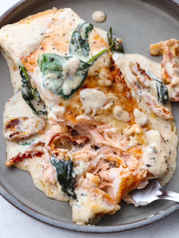 Creamy Tuscan Garlic Salmon is an easy 30 minute restaurant quality meal. The pan seared salmon gets smothered in the most amazing creamy tuscan garlic sauce with sun dried tomatoes and spinach. This is one unforgettable meal! A plate with a salmon fillet with creamy sauce and a fork cutting into it.