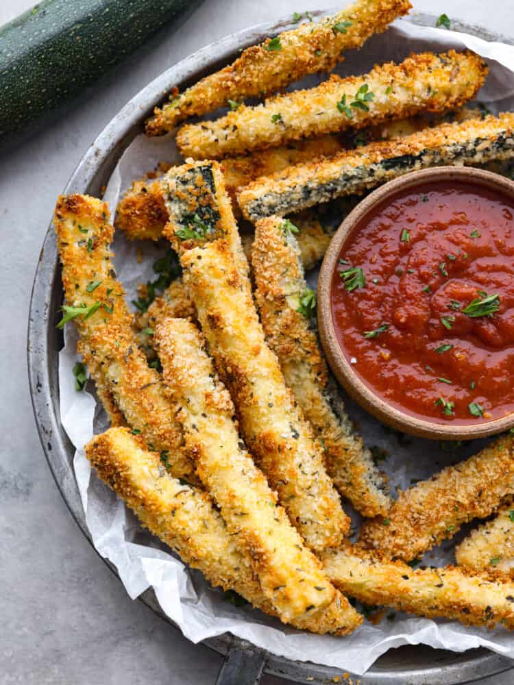 Baked zucchini fries are crispy on the outside with a crunchy, seasoned crust and tender zucchini inside. It's hard to stop after just one! Top-down view of zucchini fries served with marinara sauce on a gray plate.