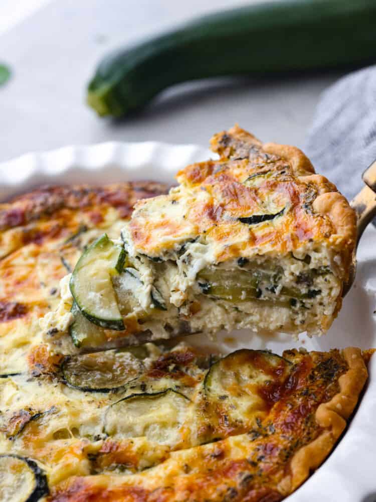 This delicious zucchini quiche has a creamy savory egg mixture baked in a perfectly flaky crust. Layers of melted cheese and zucchini make the texture of this quiche cheesy and flavorful. A slice of zucchini quiche being taken out of the serving dish with a pie server.