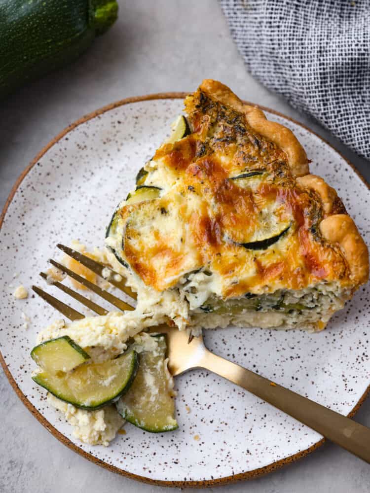 This delicious zucchini quiche has a creamy savory egg mixture baked in a perfectly flaky crust. Layers of melted cheese and zucchini make the texture of this quiche cheesy and flavorful. A slice of zucchini quiche on a plate with a fork cutting out a piece to eat.