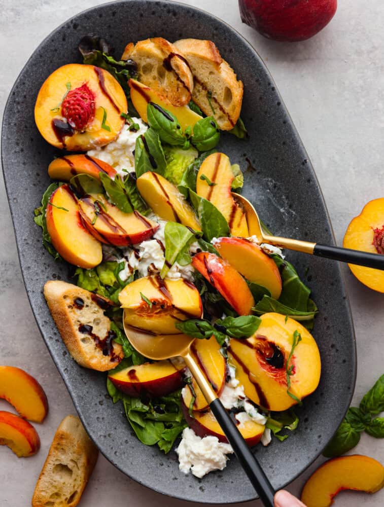 This peach burrata salad is the salad you never knew you would dream about it! Sweet ripe peaches paired with fresh creamy burrata are drizzled with balsamic glaze to marry the unique flavors all together. Top view photo of peach burrata salad on a gray platter. Black and gold serving spoons are lifting up the salad. Sliced peaches and crostini garnished on the side.