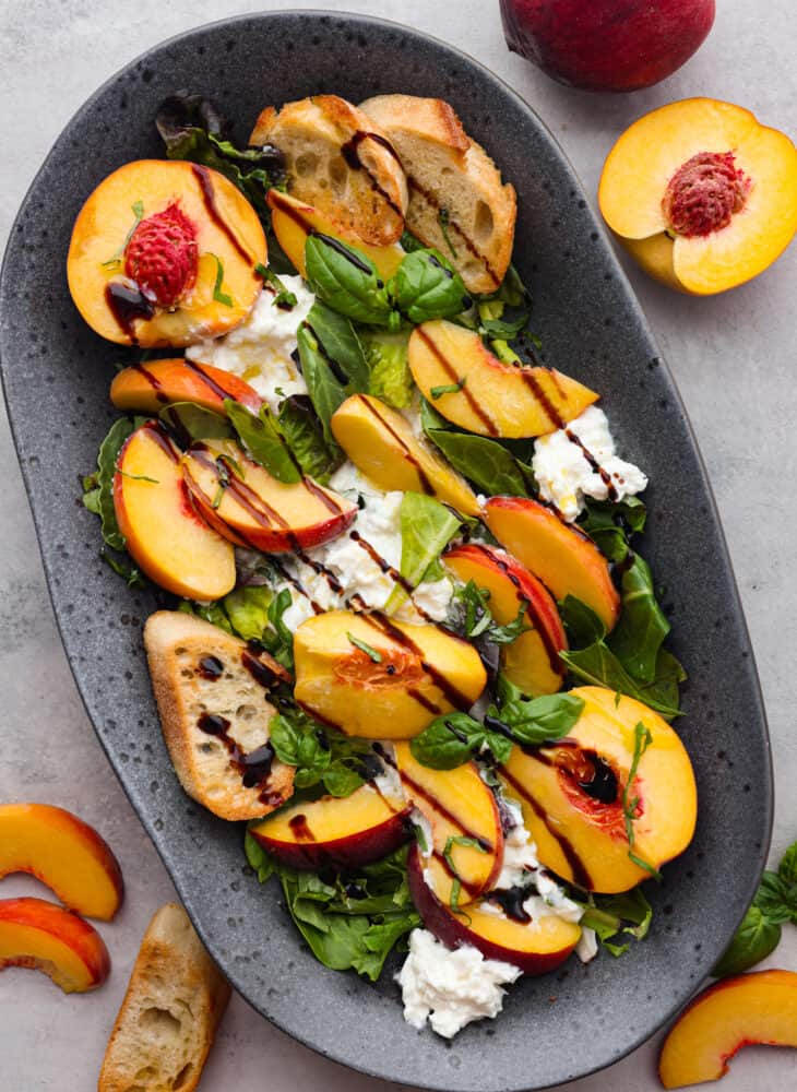 Top view photo of peach burrata salad on a gray platter. Black and gold serving spoons are lifting up the salad. Sliced peaches and crostini garnish don the side. Top view photo of peach burrata salad on a gray platter. Balsamic glaze and basil garnished on top. Sliced peaches and crostini garnished on the side. One whole and halved peach next to the platter.