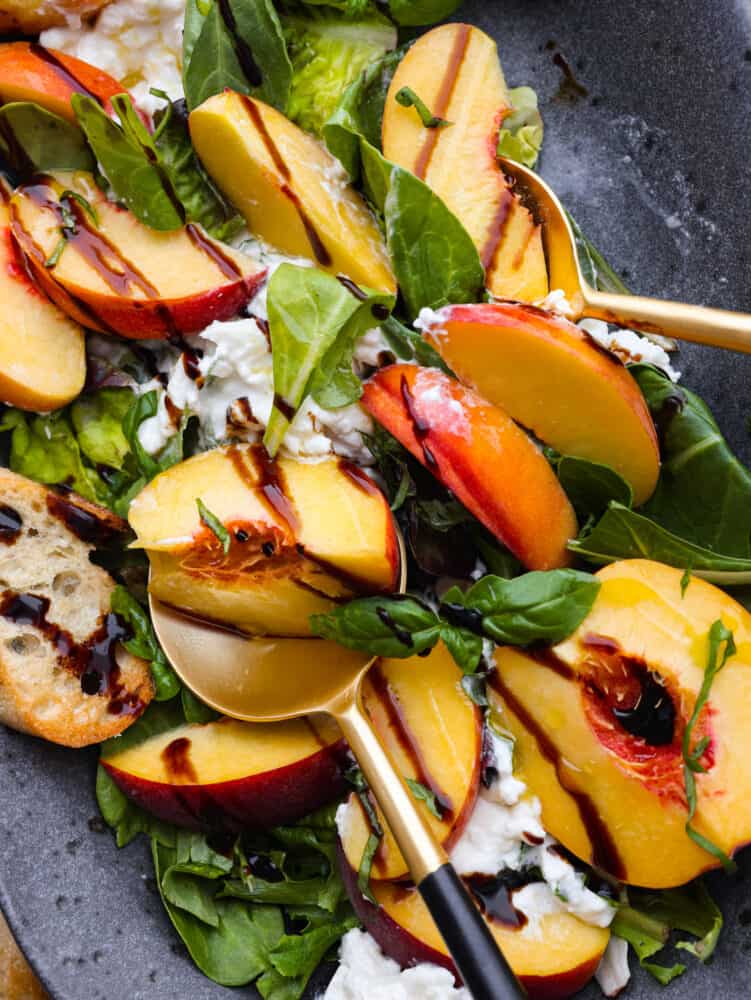 This peach burrata salad is the salad you never knew you would dream about it! Sweet ripe peaches paired with fresh creamy burrata are drizzled with balsamic glaze to marry the unique flavors all together. Close up view of peach burrata salad. Gold and black serving spoons are set in the salad. Balsamic glaze drizzled on top and garnished with crostini and fresh basil.