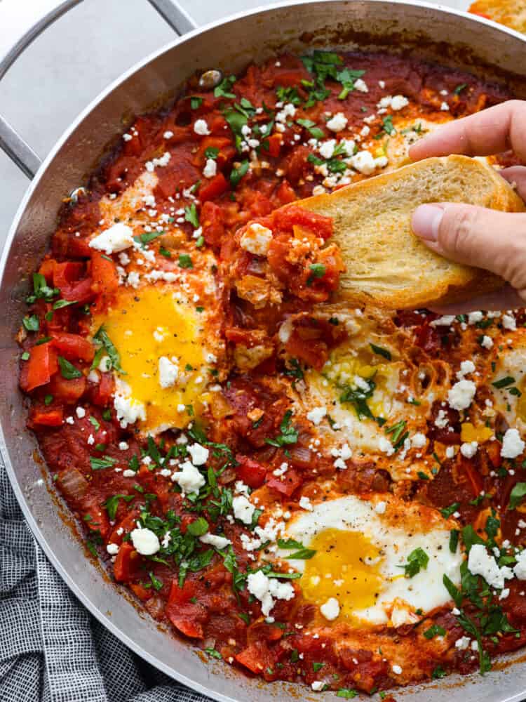 Shakshuka is an amazing one-pan meal consisting of poached eggs in a savory tomato sauce seasoned with flavorful herbs and spices! It's so simple but incredibly delicious! You'll love the bold and bright flavors inspired by North African cuisine. A piece of bread being dipped into the egg and tomato mixture.