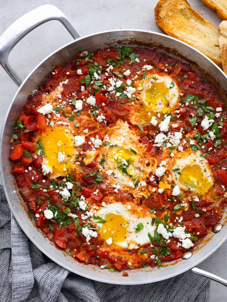 Shakshuka is an amazing one-pan meal consisting of poached eggs in a savory tomato sauce seasoned with flavorful herbs and spices! It's so simple but incredibly delicious! You'll love the bold and bright flavors inspired by North African cuisine. Top-down view of poached eggs and tomato sauce in a metal skillet.