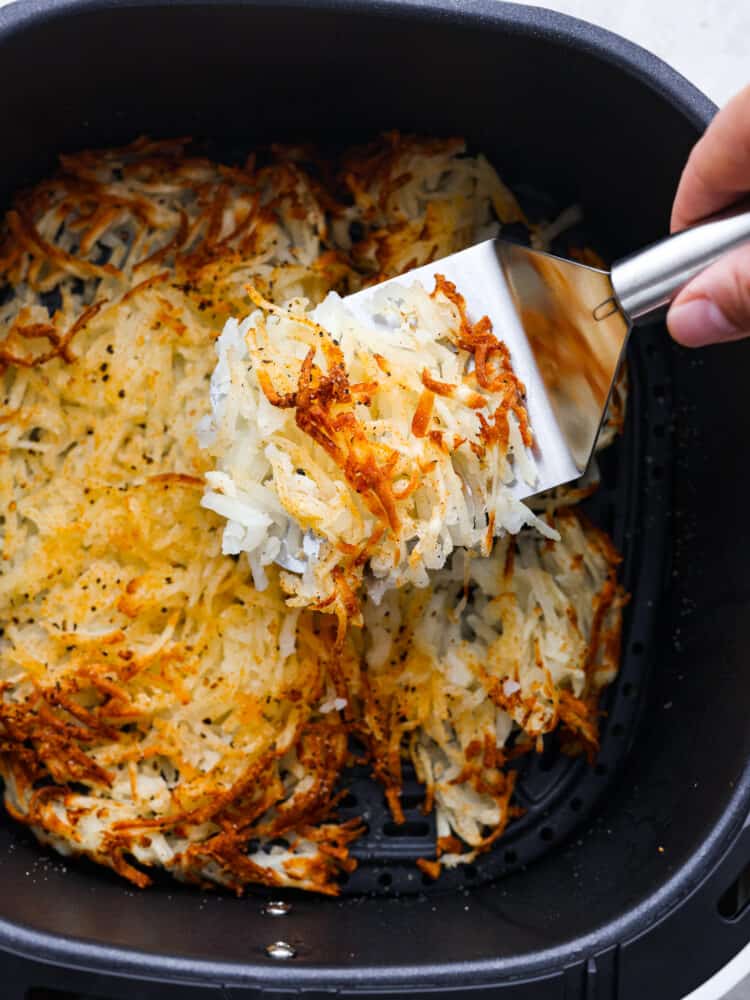 Make mornings easier with these air fryer hash browns cooked perfectly in minutes! Crispy, golden, and seasoned to taste like your favorite diner hash browns. Close up view of cooked air fryer hash browns in the air fryer. A spatula is lifting up a portion of hash browns.