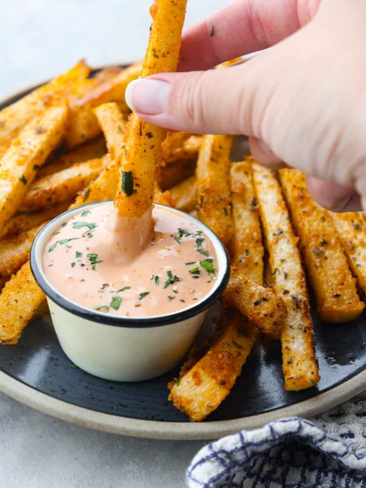 Crispy air fryer jicama fries are a surprisingly tasty and healthy alternative to fries. Enjoy seasoned low-carb fries that are both nutritious and delicious! Close up view of a jicama fry being dipped into dipping sauce on a dark gray plate. The dip is garnished with fresh parsley, and a blue and white checkered towel is next to the plate.
