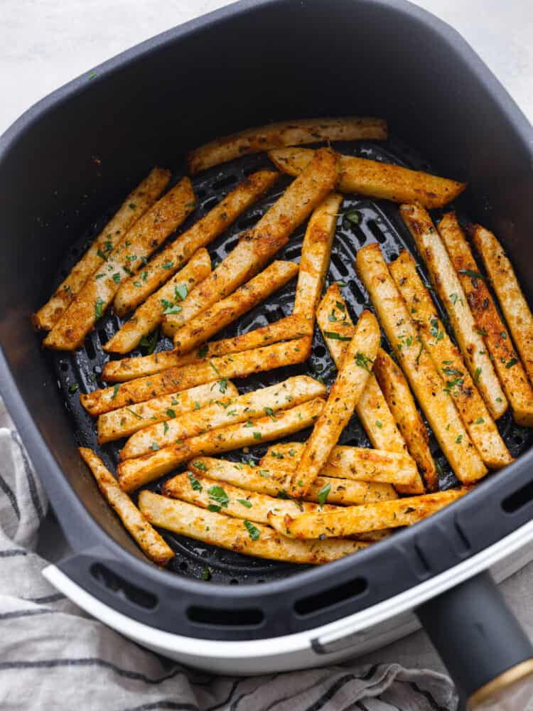 Crispy air fryer jicama fries are a surprisingly tasty and healthy alternative to fries. Enjoy seasoned low-carb fries that are both nutritious and delicious! Top view of air fryer jicama cooked in an air fryer basket. A striped hand towel is next to the basket.