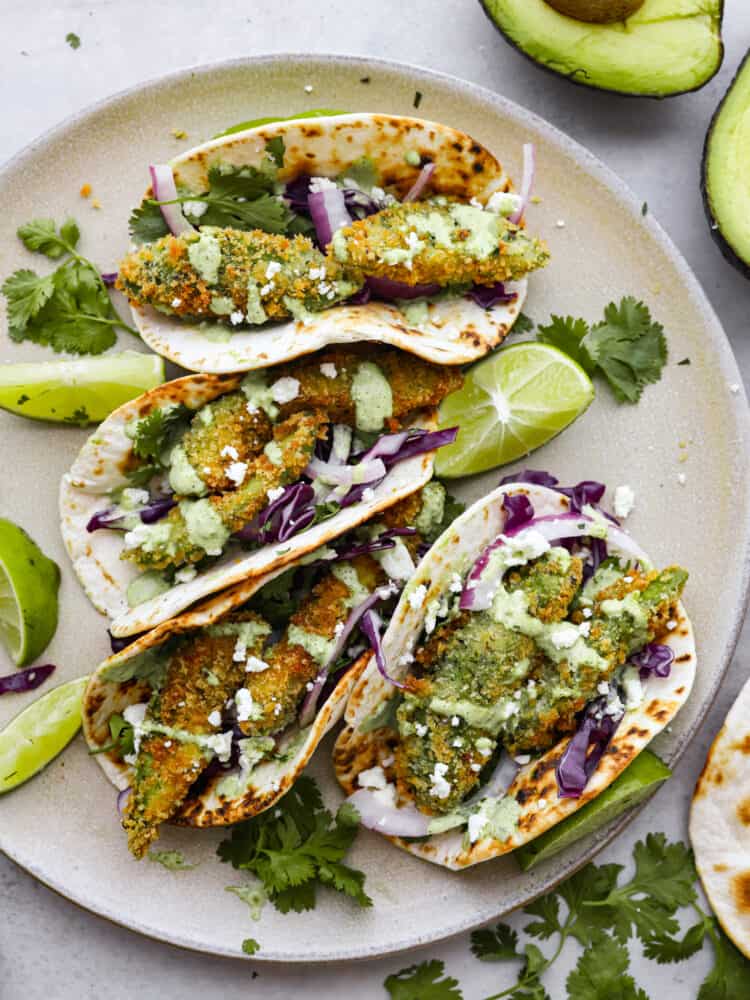 These delicious fried avocado tacos are a great option for a meat-free version of taco night! They're crispy, creamy, and topped with fresh veggies, cotija cheese, and cilantro lime dressing. Top-down view of avocado tacos arranged on a white plate.