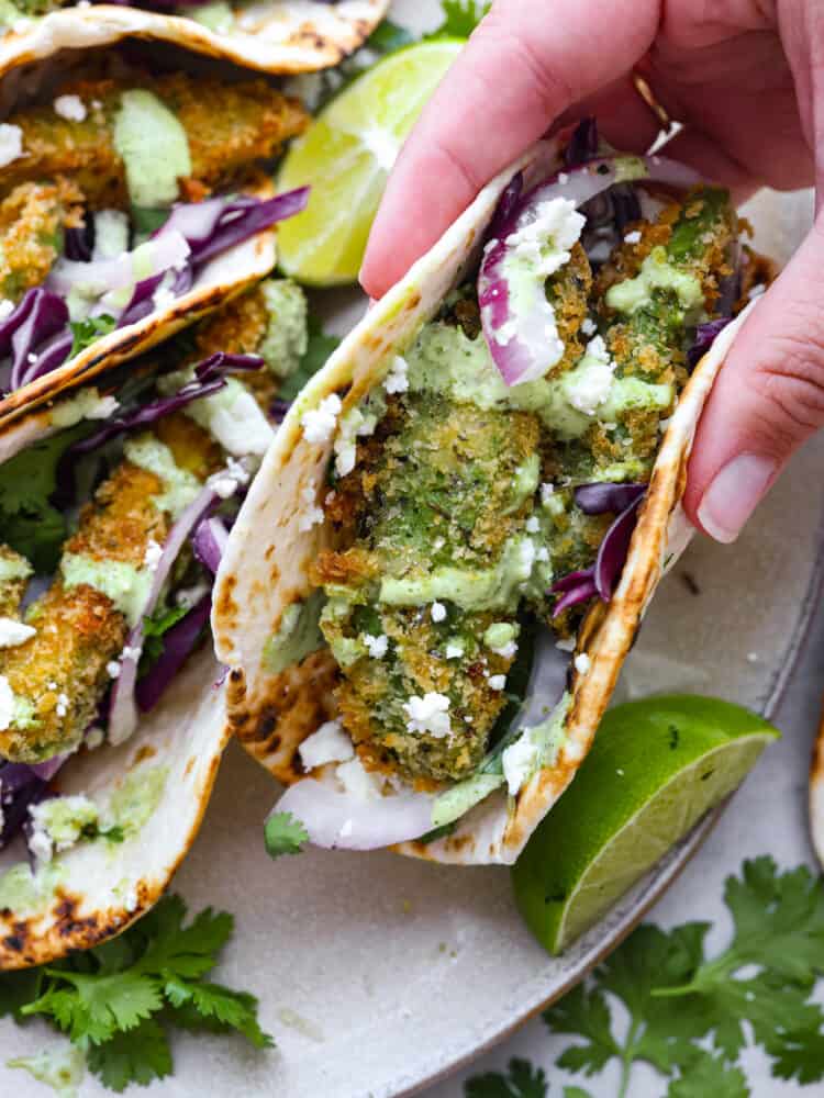 These delicious fried avocado tacos are a great option for a meat-free version of taco night! They're crispy, creamy, and topped with fresh veggies, cotija cheese, and cilantro lime dressing. A fried avocado taco being picked up.