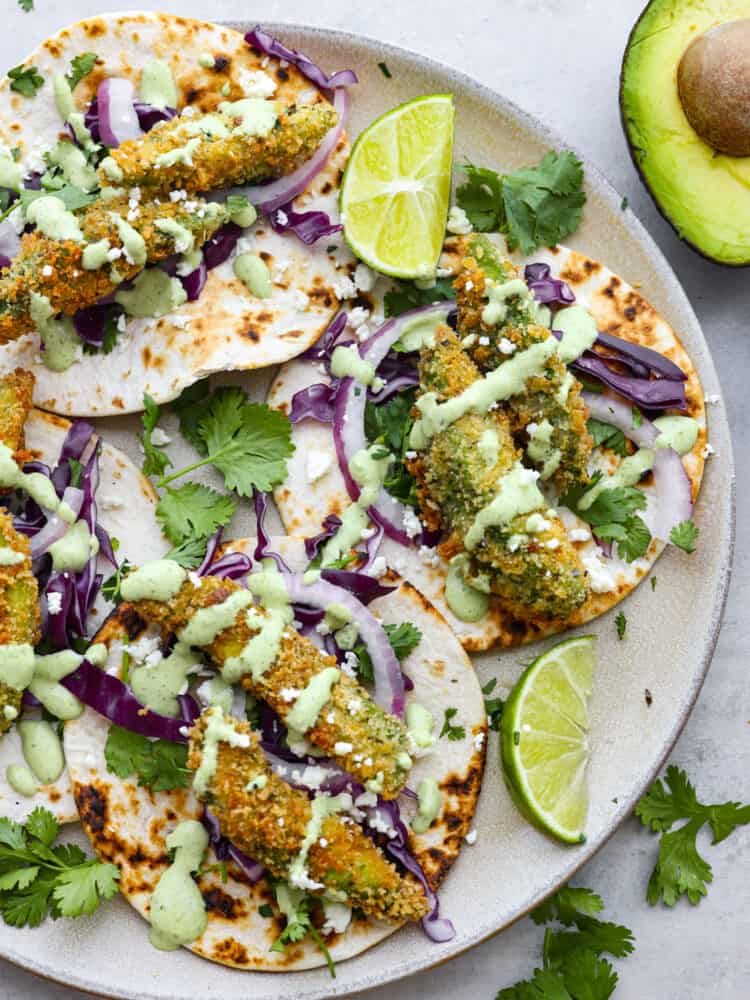 These delicious fried avocado tacos are a great option for a meat-free version of taco night! They're crispy, creamy, and topped with fresh veggies, cotija cheese, and cilantro lime dressing. 4 fried avocado tacos on a white plate.