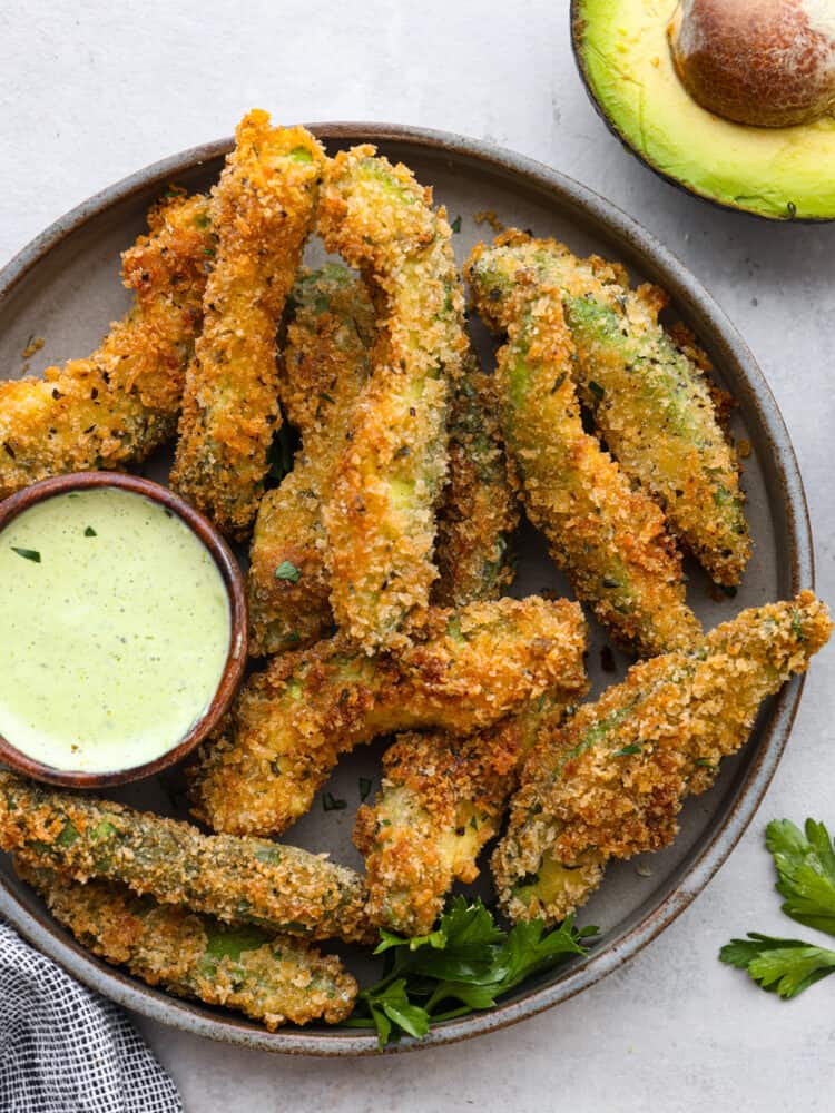 Crispy-baked avocado fries are coated in seasoning and a crunchy garlic panko crust and then baked to crispy perfection! These are so delicious and easy to make that you will want to pair them with every meal! A round platter with baked avocado fries and a small bowl filled with avocado ranch.