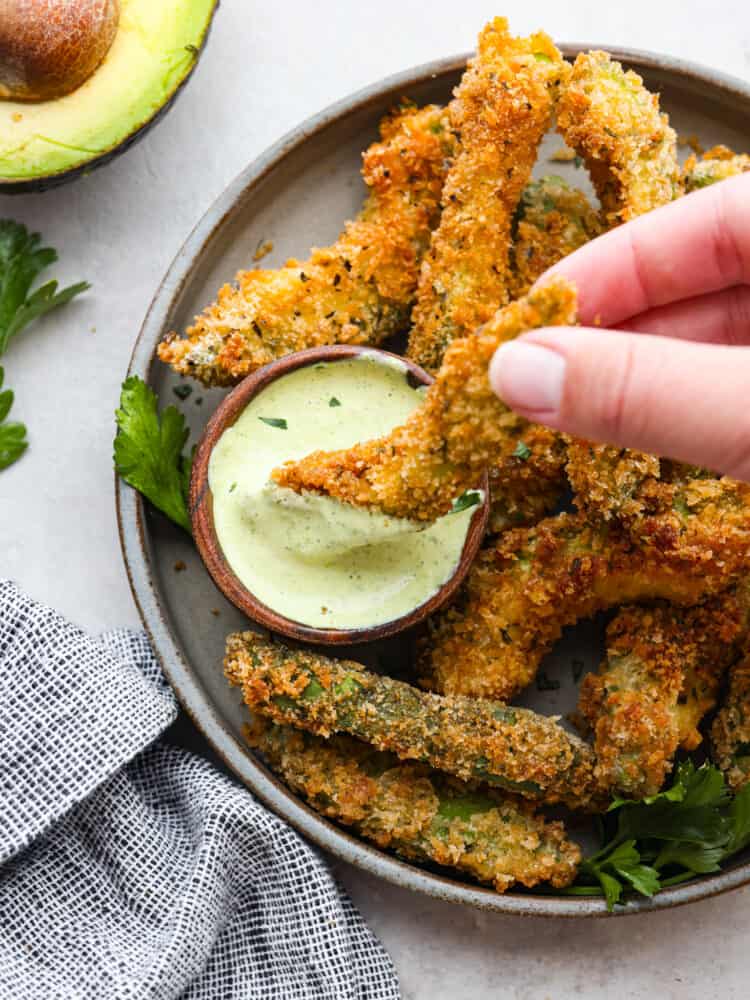 Crispy-baked avocado fries are coated in seasoning and a crunchy garlic panko crust and then baked to crispy perfection! These are so delicious and easy to make that you will want to pair them with every meal! An avocado fry being dipped into some avocado ranch.