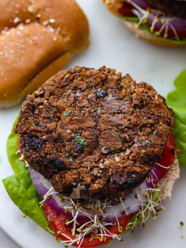 The tender meat-like texture of this black bean burger is packed with incredible flavor. Black beans blended in a mixture of veggies, flavorful herbs, and panko breadcrumbs create an amazing black bean burger that will impress everyone. Top view of a black bean burger layered on top of a bun, lettuce, red onion, tomato slice, and sprouts. The top of the bun is removed.