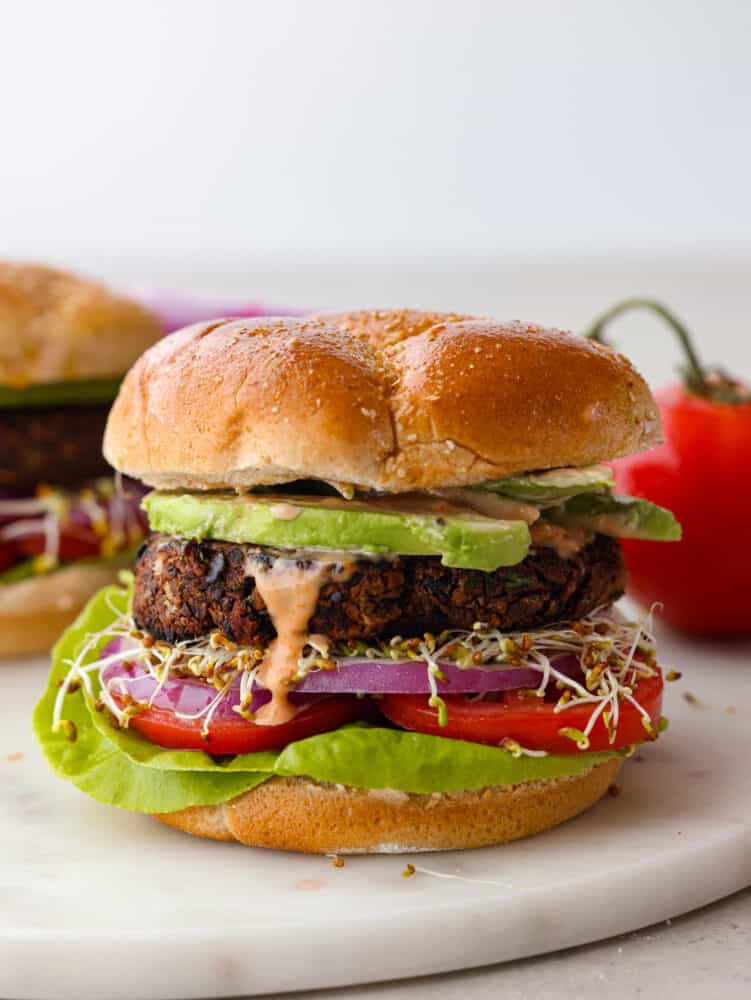 The tender meat-like texture of this black bean burger is packed with incredible flavor. Black beans blended in a mixture of veggies, flavorful herbs, and panko breadcrumbs create an amazing black bean burger that will impress everyone. A side view photo of a black bean patty layered in a bun with avocado, tomato, red onion, sprouts, lettuce, and dripping burger sauce.