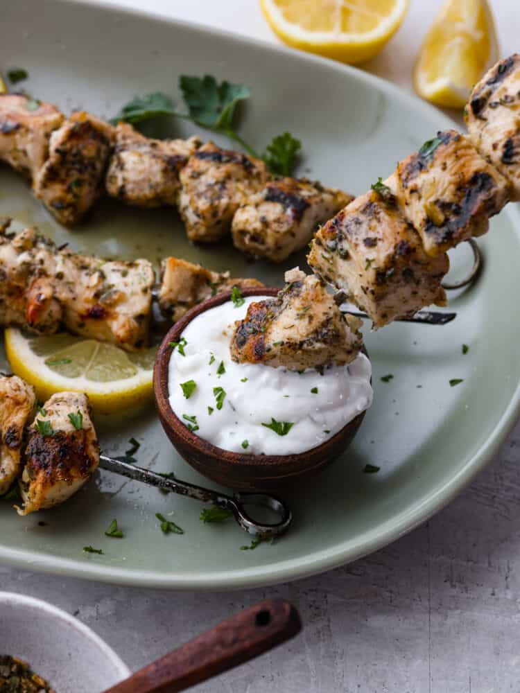 Making homemade chicken souvlaki is the best way to bring the flavors of Greek street food straight to your kitchen! These chicken skewers are juicy and coated with an amazing herb marinade. Closeup of chicken souvlaki being dipped in sauce.