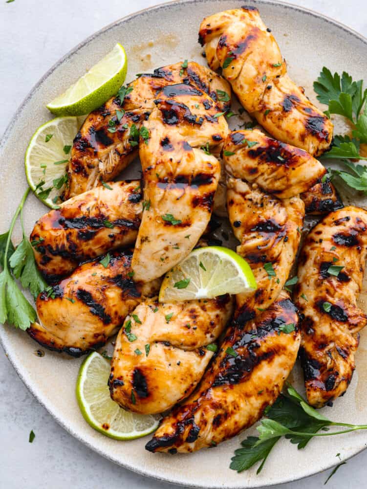 Juicy and flavorful, these 4-ingredient grilled chicken tenders are going to be your favorite thing to cook up this summer! They have a delicious honey lime glaze you'll be licking off your fingers! Top-down view of grilled chicken tenders on a gray plate, garnished with lime slices and herbs.