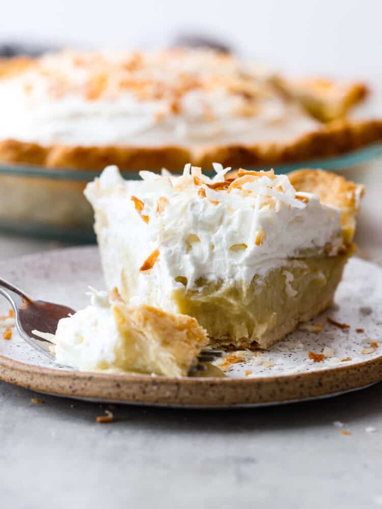 This Coconut Cream Pie recipe is made from scratch and well worth the effort! It's made with a creamy coconut custard filling and topped with sweetened whipped cream and toasted coconut. #pie #recipe #dessert #coconut A slice of coconut cream pie on a plate with a bite being cut out with a fork.