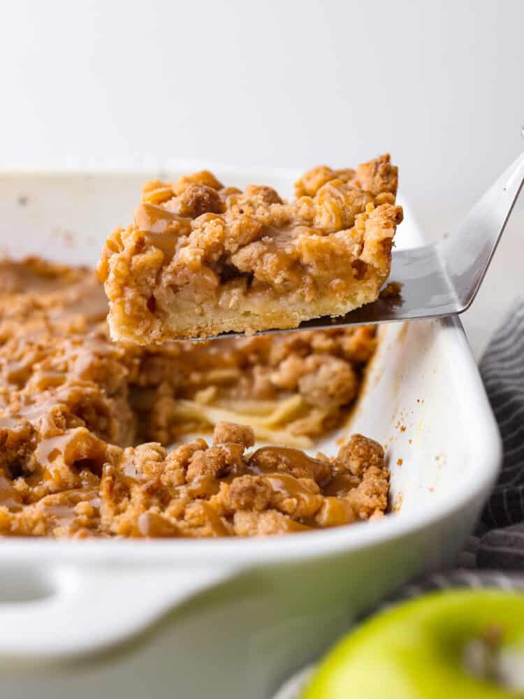 These dutch caramel apple pie bars have a flaky pie crust base with spiced apples and a buttery crumb topping. Drizzled with some homemade caramel, these will be one of the best desserts that you make this fall! A caramel apple pie bar being taken out and served from the baking dish.
