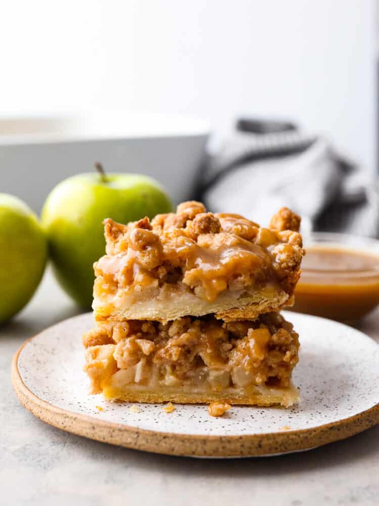 These dutch caramel apple pie bars have a flaky pie crust base with spiced apples and a buttery crumb topping. Drizzled with some homemade caramel, these will be one of the best desserts that you make this fall! A stack of 2 apple pie bars on a plate with some granny smith apples in the background.
