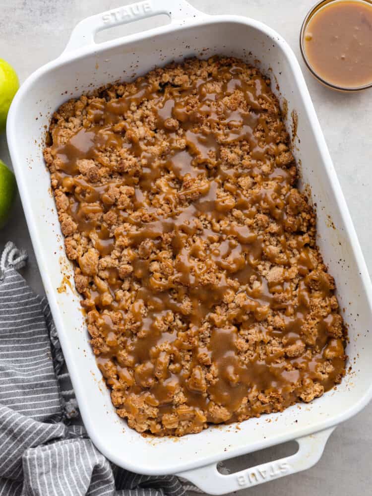 These bars have a flaky pie crust base with spiced apples and a buttery crumb topping. Drizzled with some homemade caramel, these will be one of the best desserts that you make this fall! The top view of dutch caramel apple bir bars in a baking dish, waiting to be cut.