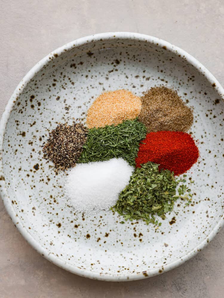 Enhance the flavor of any fish with this incredible all purpose fish seasoning. Delicious spices and herbs right from your pantry make the perfect blend to season any type of fish for grilling, baking, or pan searing. Close up photo of the seasonings and herbs clustered together on a small beige speckled plate.