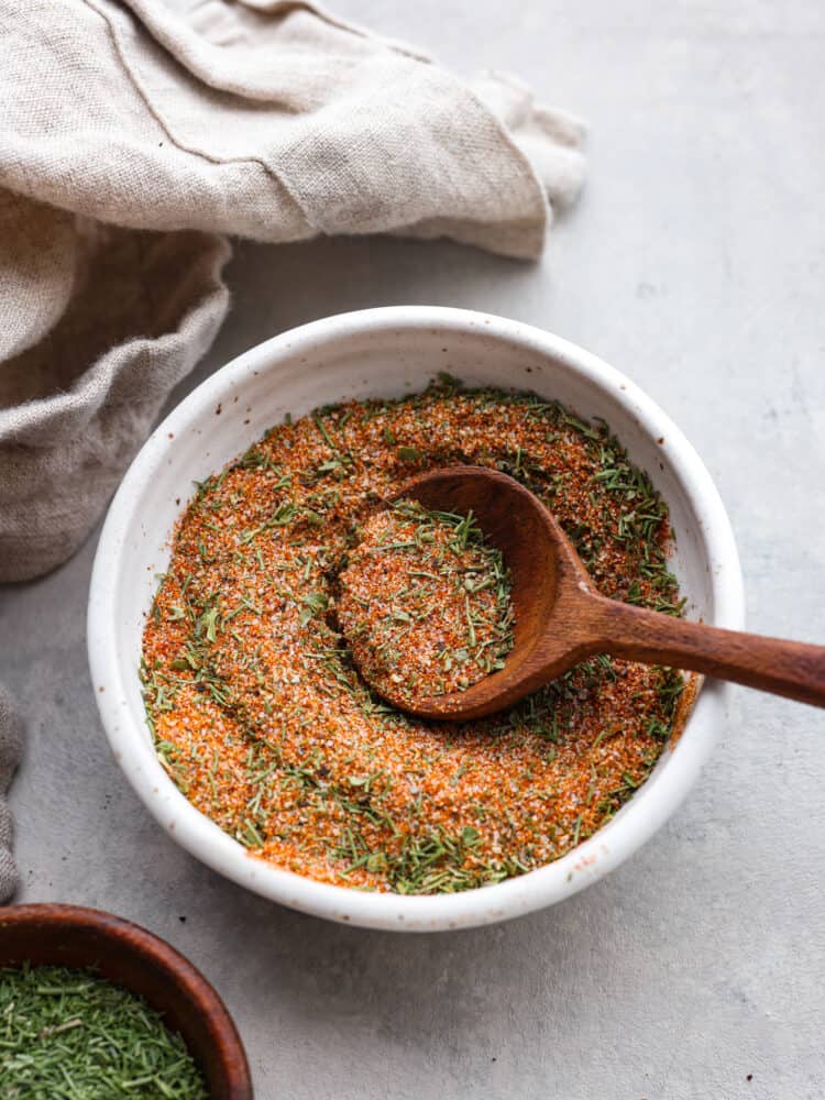 Enhance the flavor of any fish with this incredible all purpose fish seasoning. Delicious spices and herbs right from your pantry make the perfect blend to season any type of fish for grilling, baking, or pan searing. Overhead photo of fish seasoning in a cream bowl with a wood spoon scooping seasoning up. A beige bowl and wood bowl filled with dried parsley are next to the seasoning on a gray countertop.