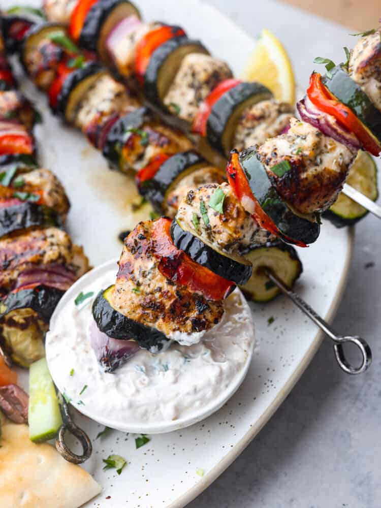 Tender Greek-flavored chicken grilled together on a skewer with fresh crisp vegetables. The Mediterranean marinade perfectly seasons these Greek chicken kabobs and transports your tastebuds to the streets of Greece. Close up photo of a Greek chicken kabob dipping in a bowl of tzatziki sauce.
