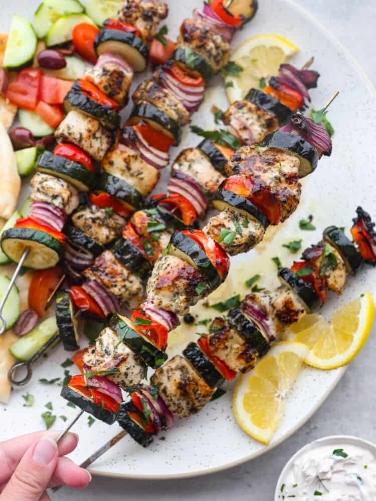 Tender Greek-flavored chicken grilled together on a skewer with fresh crisp vegetables. The Mediterranean marinade perfectly seasons these Greek chicken kabobs and transports your tastebuds to the streets of Greece. Close up photo of Greek chicken kabobs on a large white round platter. Sliced pita bread, sliced cucumbers, diced tomatoes, and kalamata olives served on the side. Sliced lemon garnished on the side with a small bowl of tzatziki sauce. A hand is holding up one of the kabobs.