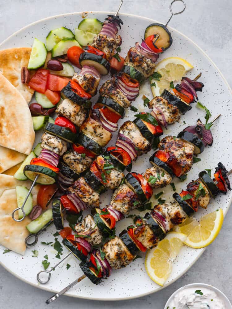 Tender Greek-flavored chicken grilled together on a skewer with fresh crisp vegetables. The Mediterranean marinade perfectly seasons these Greek chicken kabobs and transports your tastebuds to the streets of Greece. Overhead photo of Greek chicken kabobs on a large white round platter. Sliced pita bread, sliced cucumbers, diced tomatoes, and kalamata olives served on the side. Sliced lemon garnished on the side with a small bowl of tzatziki sauce.
