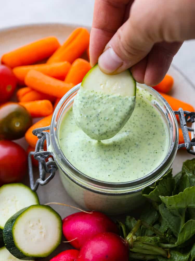 Creamy green goddess dressing is healthy and bursting with so many vibrant flavors! The combination of fresh herbs, Greek yogurt, garlic, and lemon create an irresistible dressing and dip. Top view photo of sliced zucchini being dipped into the jar of green goddess dressing. Carrots, tomatoes, zucchini, and radishes are scattered around the jar of dressing.
