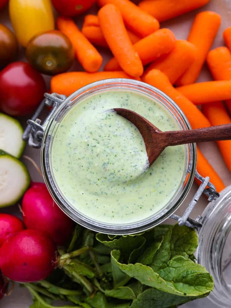 Creamy green goddess dressing is healthy and bursting with so many vibrant flavors! The combination of fresh herbs, Greek yogurt, garlic, and lemon create an irresistible dressing and dip. Close up photo of green goddess dressing in a small glass container. A wood spoon is lifting up a spoon full of dressing out of the jar. Carrots, grape tomatoes, sliced zucchini, and whole radishes scattered around the dressing.