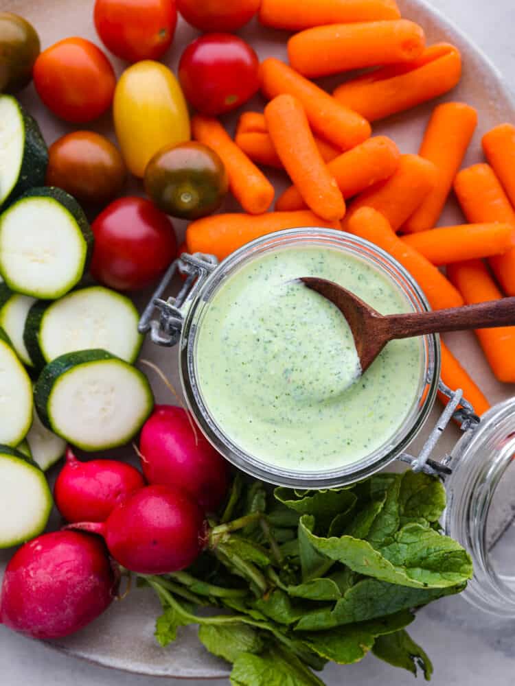Creamy green goddess dressing is healthy and bursting with so many vibrant flavors! The combination of fresh herbs, Greek yogurt, garlic, and lemon create an irresistible dressing and dip. Top view of green goddess dressing in a small glass container with a wood spoon in the jar. Carrots, grape tomatoes, sliced zucchini, and whole radishes scattered around the dressing.