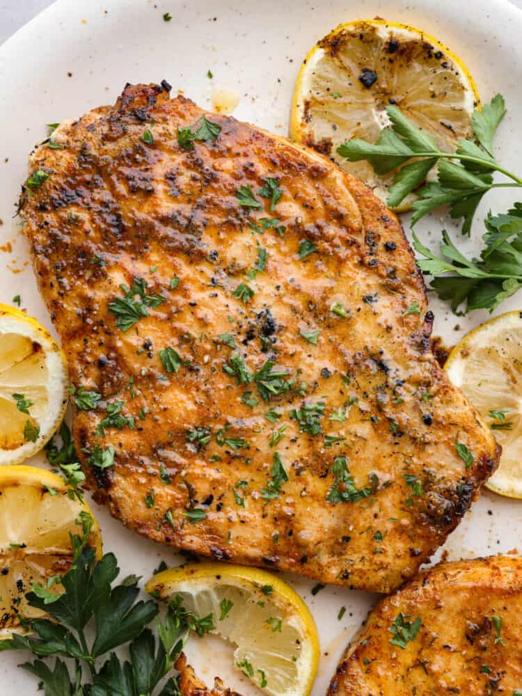 Delicious and seasoned swordfish simply grilled and seared to perfection! This tender grilled swordfish has a flavorful marinade of spices that give the fish maximum flavor. Close up view of grilled swordfish on a white platter. The swordfish is garnished with parsley and grilled lemon slices.
