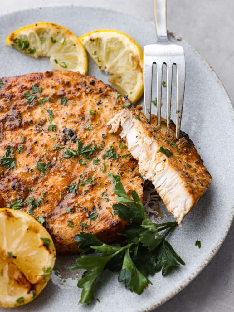 Delicious and seasoned swordfish simply grilled and seared to perfection! This tender grilled swordfish has a flavorful marinade of spices that give the fish maximum flavor. Close up photo of grilled swordfish with a fork lifting up a cut piece. Garnished with grilled lemon wedges and parsley leaves.