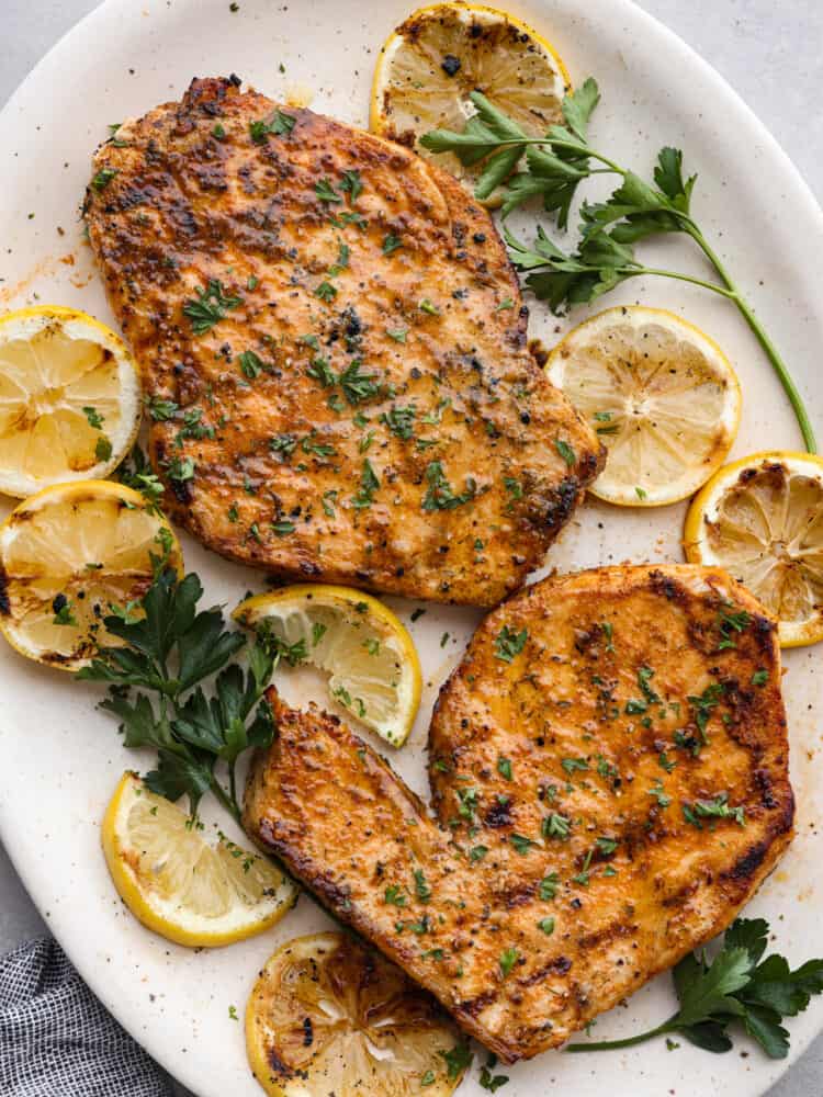 Delicious and seasoned swordfish simply grilled and seared to perfection! This tender grilled swordfish has a flavorful marinade of spices that give the fish maximum flavor. Overhead view of two grilled swordfish steaks on a large white platter. Grilled lemon slices and parsley garnished on the side.