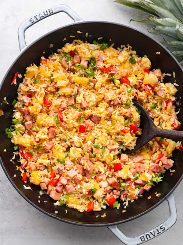 This Hawaiian fried rice is loaded with sweet pineapple, ham, and veggies and is so much better than takeout! Everyone goes crazy over this meal, and it's definitely a family favorite! The top view of Hawaiian fried rice cooked in a skillet with a wooden spoon.