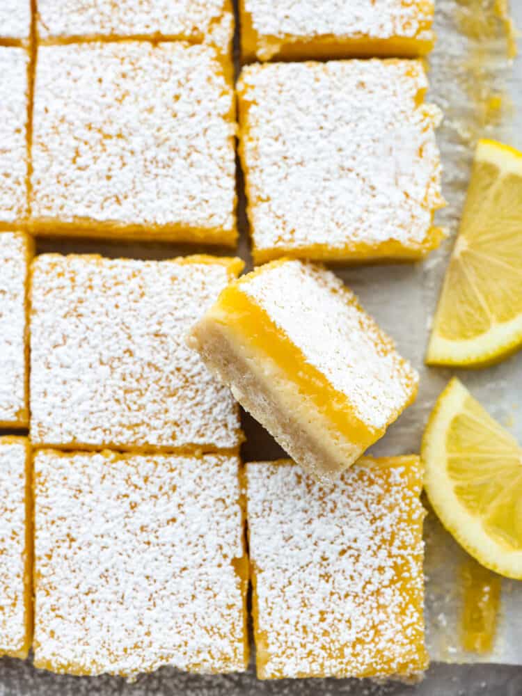 These are the most delicious and creamy lemon cheesecake bars. They have a buttery shortbread crust with a cheesecake and lemon layer and are dusted with powdered sugar. They are the perfect dessert! Lemon cheesecake bars on parchment paper. One is turned to the side so that you can see all of the layers.